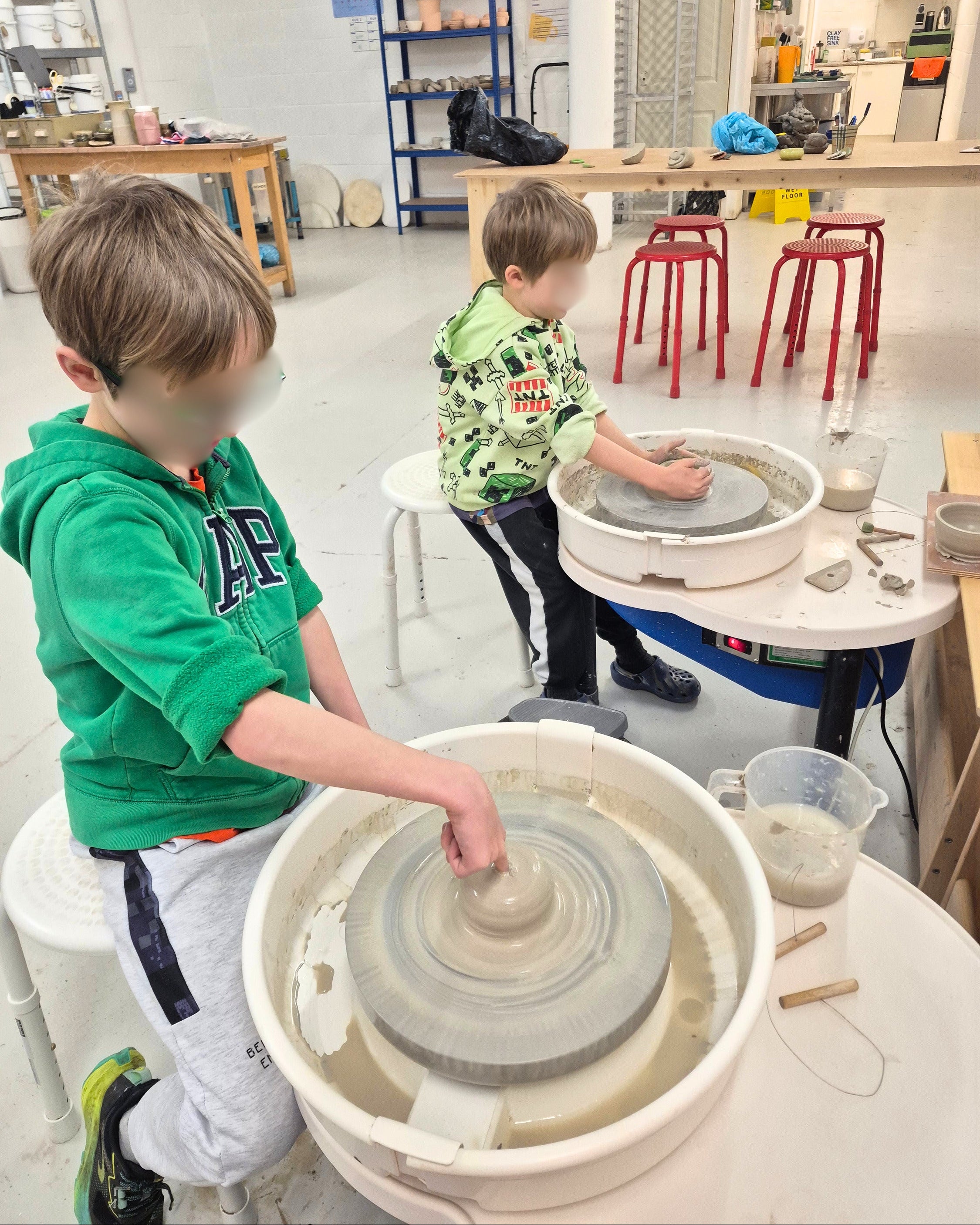 Two children practicing pottery in a studio, each working at a spinning wheel with clay. One child in a green hoodie is in the foreground, and another child in a green camouflage shirt is seated further back.