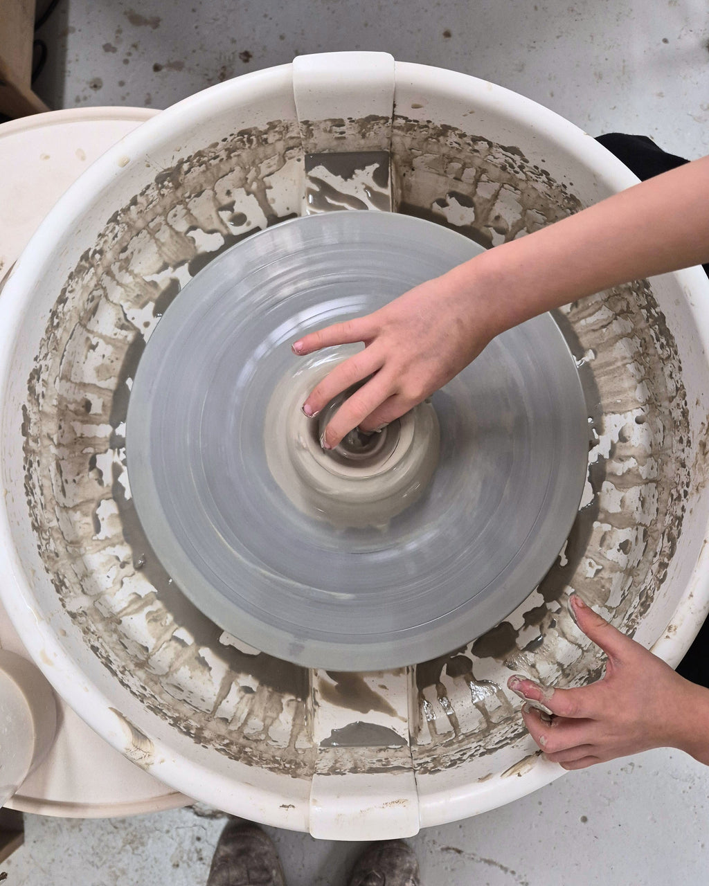 A top-down view of a pottery wheel in motion, with a child's hands guiding a small lump of clay into a spinning wheel. The white wheel is covered in wet clay residue. 
