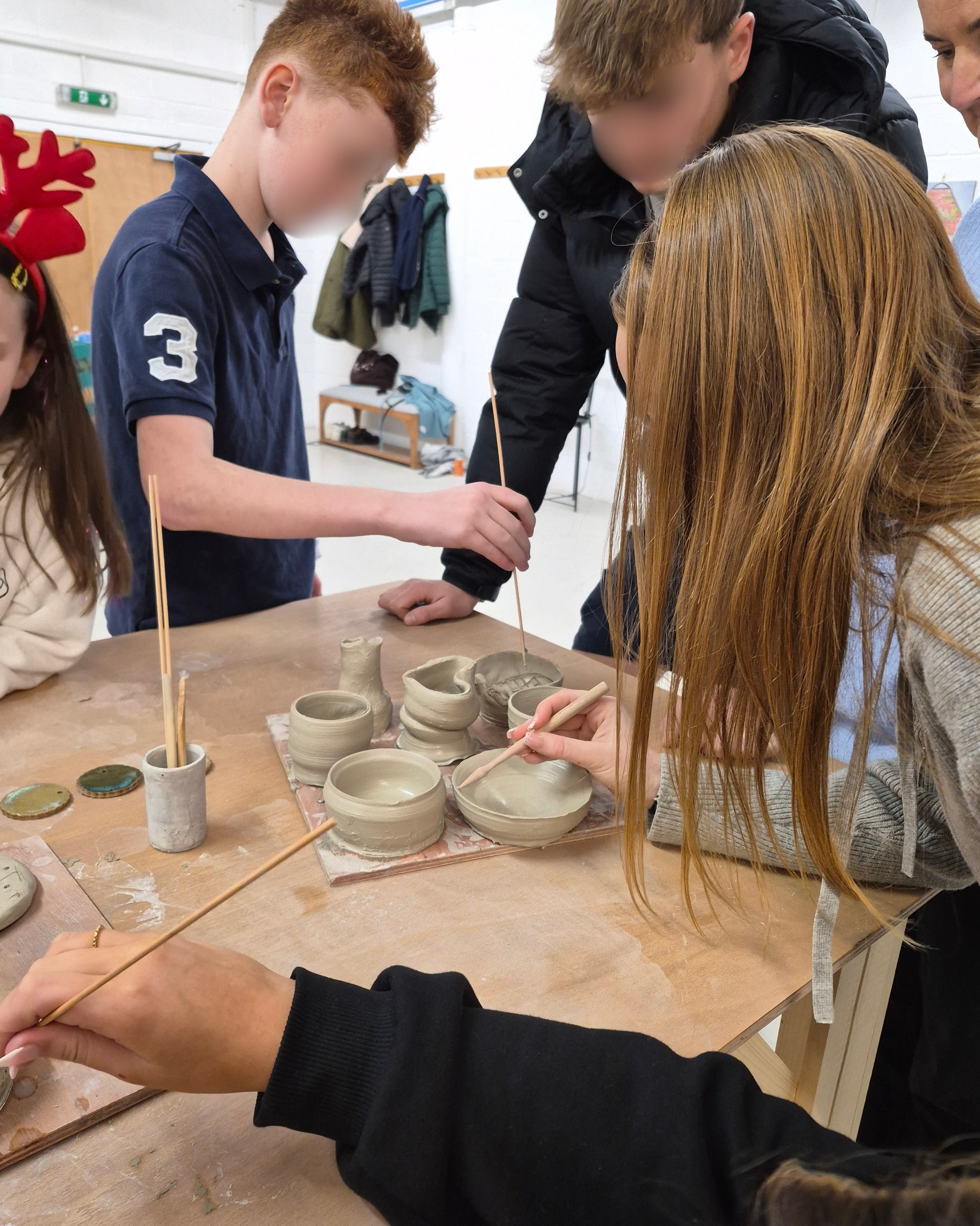 Teenagers working together at a pottery table, shaping small clay pots and vessels with hand tools on a wooden table in a bright indoor studio.