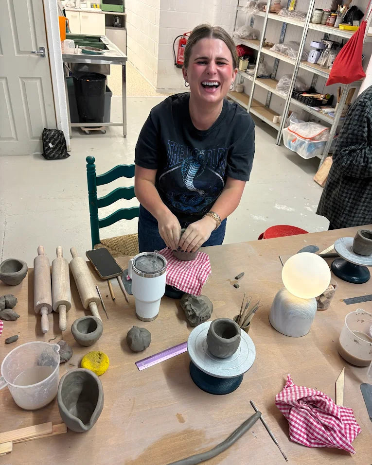 Person sitting at a table with pottery-making tools and clay items in a workshop.